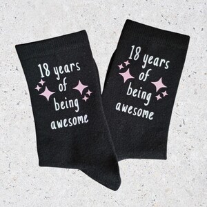 Op de afbeelding: Een paar zwarte sokken met de tekst "18 years of being awesome" in het wit en roze sterren. Een feestelijk cadeau voor een 18e verjaardag.