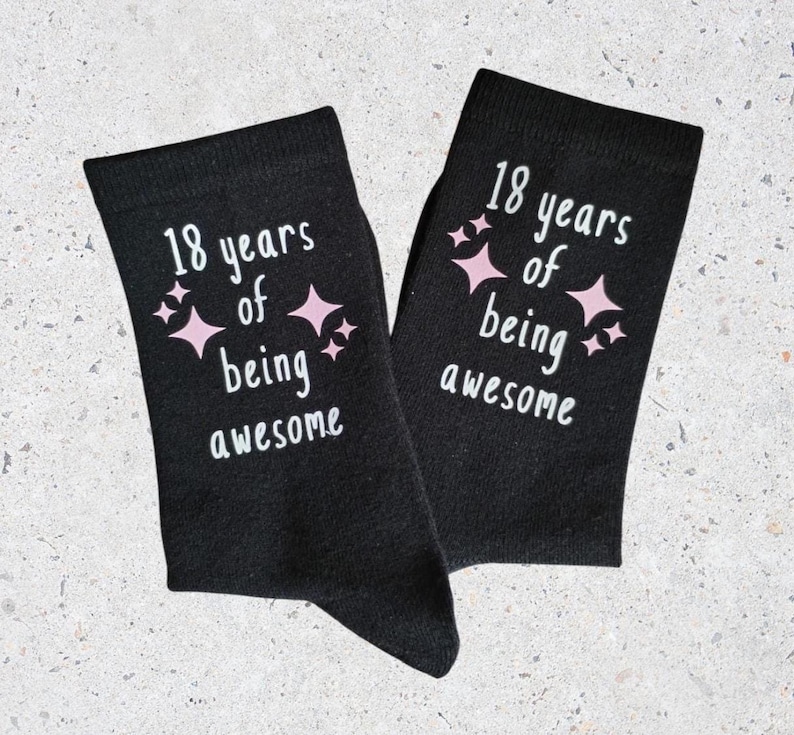 Op de afbeelding: Een paar zwarte sokken met de tekst "18 years of being awesome" in het wit en roze sterren. Een feestelijk cadeau voor een 18e verjaardag.