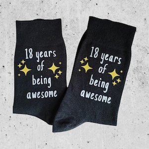 Op de afbeelding: Een paar zwarte sokken met de tekst "18 years of being awesome" in het wit, versierd met gele sterren. De sokken worden gepresenteerd op een lichtgrijze ondergrond.