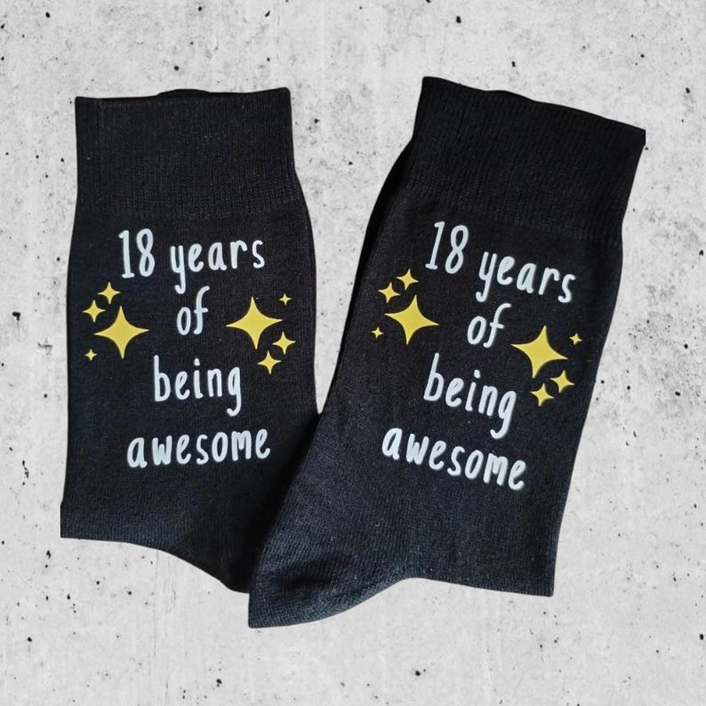 Op de afbeelding: Een paar zwarte sokken met de tekst "18 years of being awesome" in het wit, versierd met gele sterren. De sokken worden gepresenteerd op een lichtgrijze ondergrond.