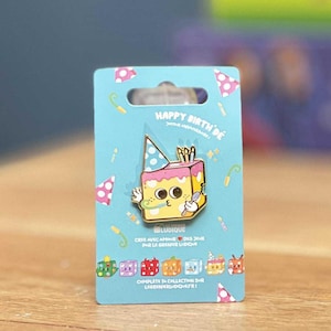 Peut inclure: Épingle en émail doré représentant un gâteau de dessin animé avec un chapeau de fête, des bougies et un couteau, sur une carte bleue. La carte porte le texte "HAPPY BIRTH DE" et "LUDIQUE". L'épingle est un objet de collection.