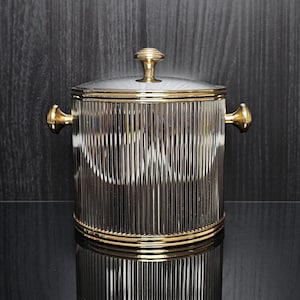 Op de afbeelding: Een heldere glazen ijsemmer met verticale geribbelde details en goudkleurige accenten. De emmer heeft een deksel met een gouden knop en twee gouden handvatten. De ijsemmer staat op een reflecterend zwart oppervlak.