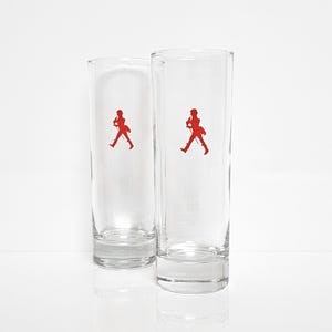 Peut inclure: Deux verres &agrave; boire cylindriques transparents avec un logo rouge repr&eacute;sentant une silhouette d'homme en marche. Les verres sont en verre transparent et sont pos&eacute;s sur un fond blanc.