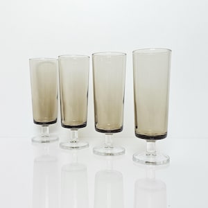 Op de afbeelding: Een set van vier hoge, slanke drinkglazen met een rookgrijze tint. Elk glas heeft een cilindrische kelk die iets taps toeloopt naar de basis, ondersteund door een korte, heldere steel en een ronde voet, wat zorgt voor een uniform uiterlijk.