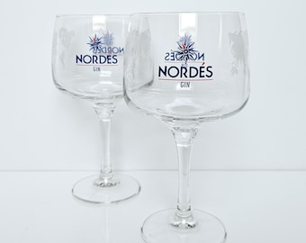 Könnte beinhalten: Zwei klare Gin-Gläser mit dem Wort "NORDÉS" in Blau und Weiß aufgedruckt. Die Gläser haben eine breite Schale und einen schlanken Stiel. Der Hintergrund ist schlicht weiß.