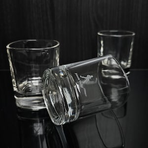 May include: Three clear, square-shaped shot glasses are arranged on a reflective black surface. One glass is lying on its side, displaying an etched design. The other two glasses are upright, with the background a dark wood.