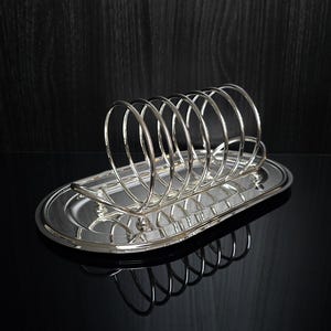 Puede incluir: Tostador de metal plateado con divisores de alambre curvos, colocado sobre una bandeja ovalada. El soporte está diseñado para contener varias rebanadas de tostadas. La superficie reflectante crea un efecto de espejo.