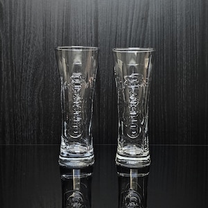 Könnte beinhalten: Zwei klare, hohe Trinkgläser mit geätzten Designs. Die Gläser sind zylindrisch mit leicht ausgestelltem Oberteil und stehen auf einer reflektierenden schwarzen Oberfläche. Der Hintergrund ist dunkles Holzmaserung.