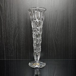 Op de afbeelding: Een heldere, geslepen glazen vaas met een uitlopende bovenkant en een lange, getextureerde body. De vaas heeft een ronde basis en is ongeveer 25 cm hoog. Het ontwerp heeft een herhalend patroon van ruitvormige sneden. De vaas staat op een reflecterend oppervlak.