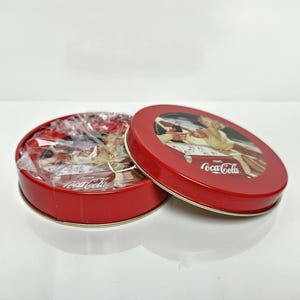 Può includere: Una scatola rotonda rossa della Coca-Cola con un coperchio aperto, che rivela caramelle confezionate. Il coperchio presenta una pubblicità vintage della Coca-Cola. La scatola è su una superficie bianca. Il logo Coca-Cola è visibile.