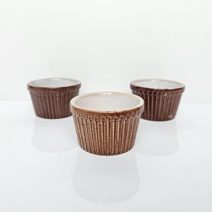 Op de afbeelding: Drie kleine, ronde keramische bakjes met een bruine, verticaal geribbelde buitenkant en een gladde, witte binnenkant. De schaaltjes zijn in een driehoeksvorm geplaatst en tonen hun consistente ontwerp, uitlopende randen en glanzende afwerking.