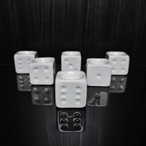 Peut inclure: Un ensemble de cinq coquetiers en céramique blanche en forme de dés à six faces. Chaque cube présente des points circulaires en creux sur les côtés et une dépression circulaire sur le dessus pour maintenir un œuf. Les dés sont disposés en triangle.