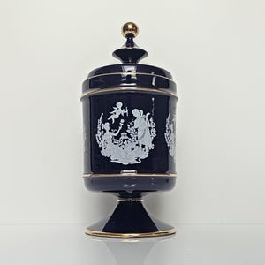 Op de afbeelding: Een donkerblauwe glazen pot met bijpassend deksel en een goudkleurige knop. Het cilindrische lichaam is versierd met witte reliëffiguren in camee-stijl van cherubijnen en klassieke figuren, geaccentueerd door een dunne gouden rand langs de randen.