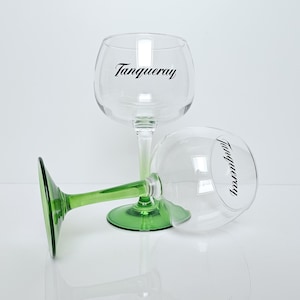 Op de afbeelding: Twee heldere gin-glazen met groene stelen en voet, één rechtop en één op zijn kant. Het woord "Tanqueray" is in het zwart op het glas gedrukt. De glazen staan op een witte ondergrond.