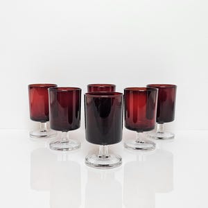 Op de afbeelding: Een set van zes robijnrode wijnglazen. De glazen hebben een cilindrische kelk en een heldere, korte steel. De glazen zijn gerangschikt op een wit oppervlak, met hun reflecties zichtbaar. De glazen zijn ongeveer 10 cm hoog.