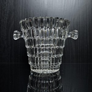 Op de afbeelding: Een heldere, facetgeslepen glazen ijsemmer met twee sierlijke handvatten. De emmer heeft een gestructureerd, geometrisch ontwerp en staat op een reflecterend oppervlak, waardoor een spiegeleffect ontstaat. De achtergrond is donker.