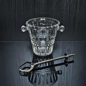 Op de afbeelding: Een heldere, gefacetteerde glazen ijsemmer met twee handvatten aan de zijkant en een geometrisch patroon, vergezeld van een lange, slanke zilverkleurige metalen ijstang met een veer-mechanisme en een klein stervormig detail aan het uiteinde.