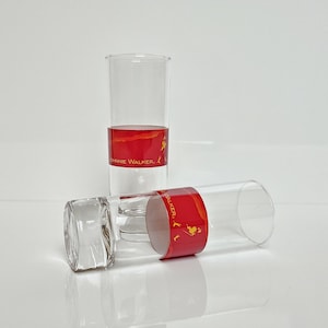 May include: Two clear, cylindrical drinking glasses with a red band featuring gold text and a logo. One glass stands upright, the other lies on its side. The glasses are on a white surface.