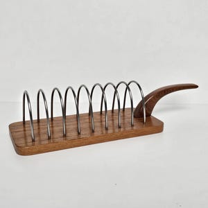 May include: A rectangular wooden toast rack featuring eight arched metal wire dividers and a curved wooden handle extending from one side. The base is made of polished brown wood with a visible grain, and the metal arches are dark grey in color.