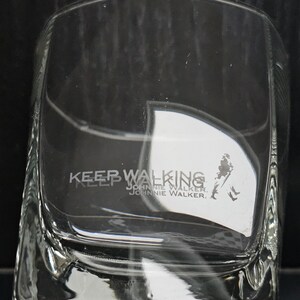May include: Clear, square-shaped glass with the words "KEEP WALKING" and "JOHNNIE WALKER" etched on the front. A silhouette of a man walking is also visible. The glass is designed for spirits.