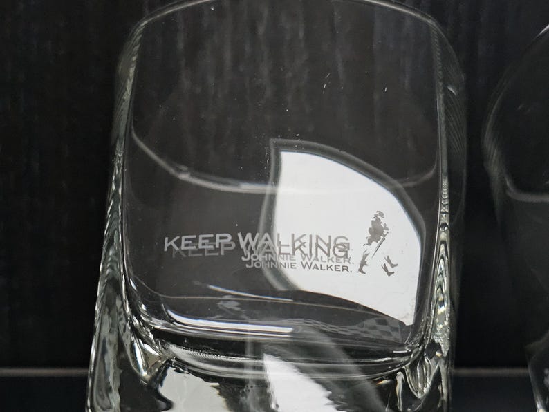 May include: Clear, square-shaped glass with the words "KEEP WALKING" and "JOHNNIE WALKER" etched on the front. A silhouette of a man walking is also visible. The glass is designed for spirits.