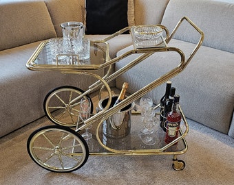 Op de afbeelding: Een gouden barwagen met twee glazen planken, een zilveren ijsbak en flessen wijn en sterke drank. De wagen heeft vier wielen en staat op een lichtgrijs tapijt.