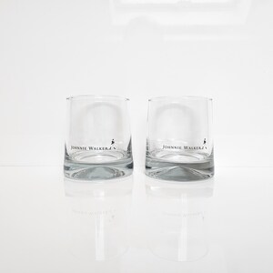 Peut inclure: Deux verres &agrave; whisky bas et transparents avec le logo "Johnnie Walker" imprim&eacute; sur le devant. Les verres ont une base &eacute;paisse et incurv&eacute;e et se refl&egrave;tent sur une surface blanche. Le logo pr&eacute;sente le nom de la marque et une silhouette d'homme.