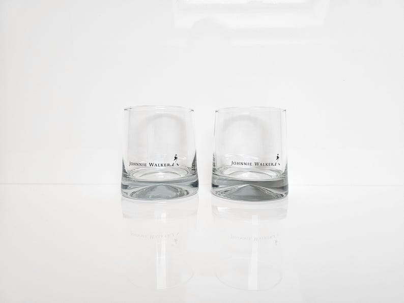 Peut inclure: Deux verres &agrave; whisky bas et transparents avec le logo "Johnnie Walker" imprim&eacute; sur le devant. Les verres ont une base &eacute;paisse et incurv&eacute;e et se refl&egrave;tent sur une surface blanche. Le logo pr&eacute;sente le nom de la marque et une silhouette d'homme.