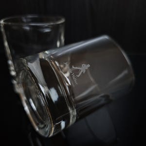 May include: Two clear, lowball whiskey glasses with the Johnnie Walker logo etched on the side. One glass is upright, the other is tilted, with a dark background. The logo features a walking figure.