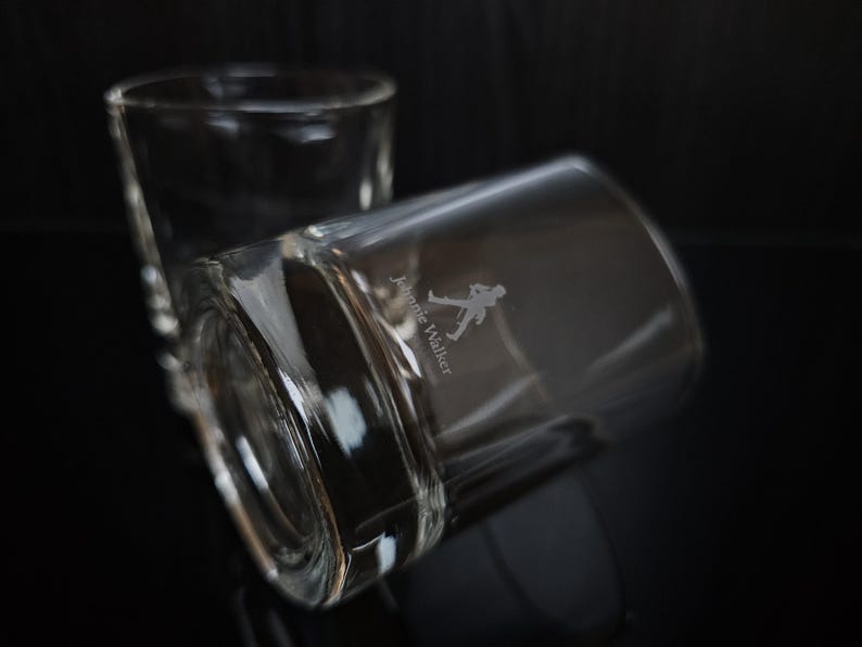 May include: Two clear, lowball whiskey glasses with the Johnnie Walker logo etched on the side. One glass is upright, the other is tilted, with a dark background. The logo features a walking figure.