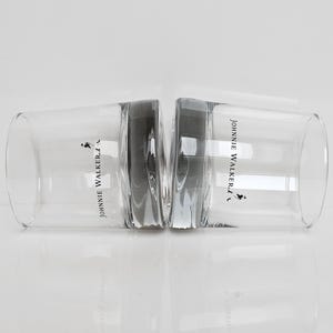 Peut inclure: Deux verres &agrave; whisky bas et transparents avec le logo "Johnnie Walker" et un homme en marche. Les verres sont c&ocirc;te &agrave; c&ocirc;te, se refl&eacute;tant sur une surface blanche. Les verres sont en verre transparent.