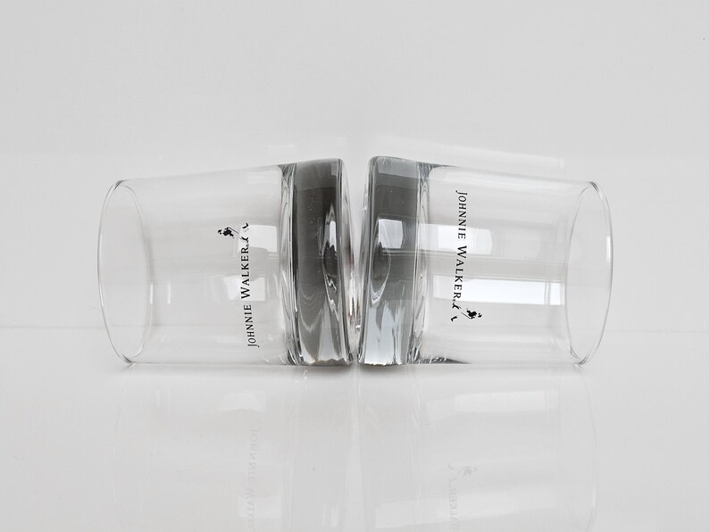 Peut inclure: Deux verres &agrave; whisky bas et transparents avec le logo "Johnnie Walker" et un homme en marche. Les verres sont c&ocirc;te &agrave; c&ocirc;te, se refl&eacute;tant sur une surface blanche. Les verres sont en verre transparent.