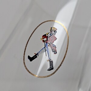 May include: A clear glass bottle with a gold oval border. Inside the border is a colorful illustration of a person walking with a cane. The person is wearing a red coat, blue pants, and black boots.