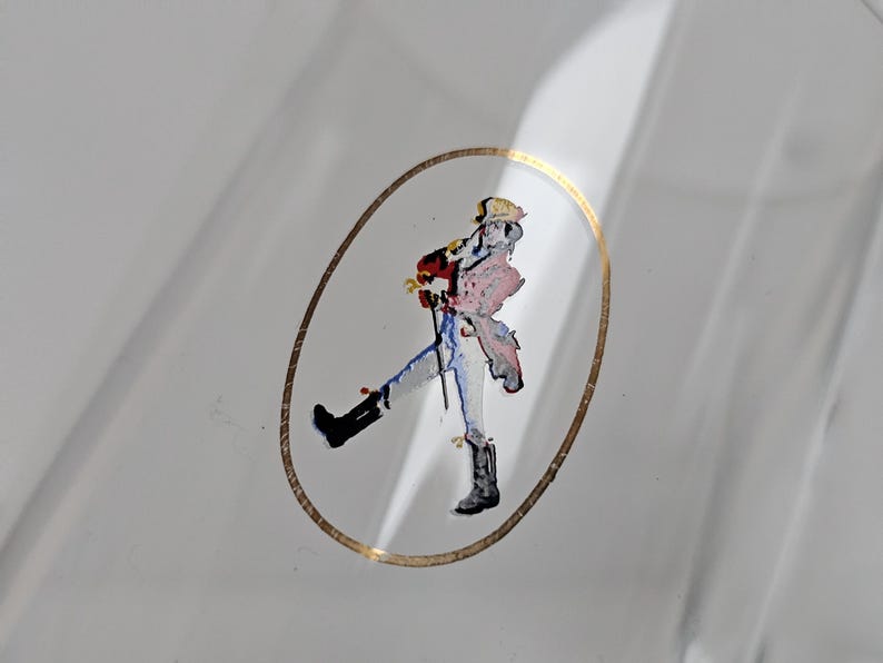 May include: A clear glass bottle with a gold oval border. Inside the border is a colorful illustration of a person walking with a cane. The person is wearing a red coat, blue pants, and black boots.