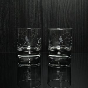 May include: Two clear, lowball whiskey glasses with the Johnnie Walker logo etched on the front. The logo features a silhouette of a man walking. The glasses are reflected on a black surface.