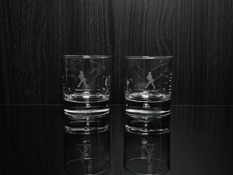 May include: Two clear, lowball whiskey glasses with the Johnnie Walker logo etched on the front. The logo features a silhouette of a man walking. The glasses are reflected on a black surface.