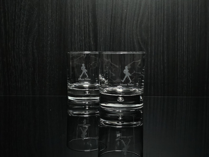 May include: Two clear, lowball whiskey glasses with the Johnnie Walker logo etched on the front. The logo features a silhouette of a man walking. The glasses are reflected on a glossy black surface.