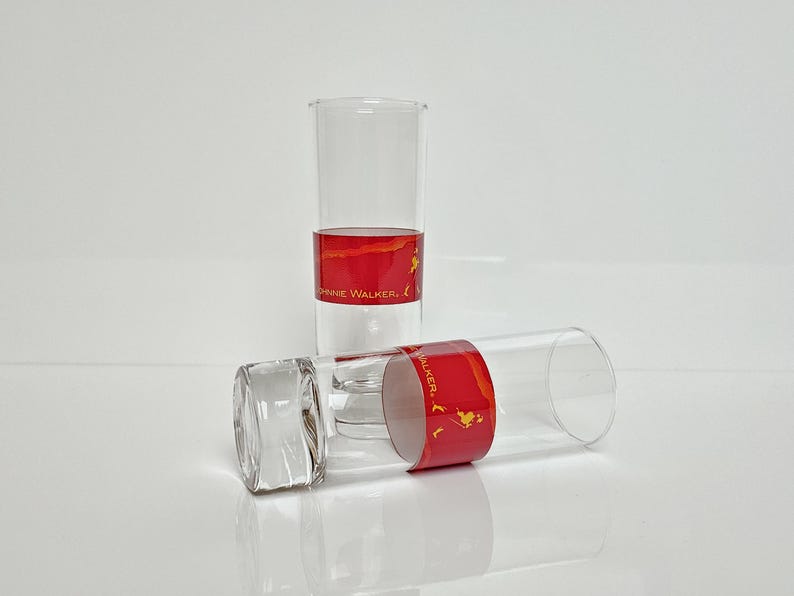May include: Two clear, cylindrical drinking glasses with a red band featuring gold text and a logo. One glass stands upright, the other lies on its side. The glasses are on a white surface.