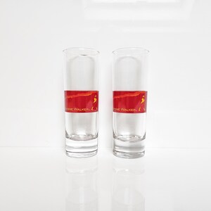 May include: Two clear, tall shot glasses with a red band featuring the text "Johnnie Walker". The glasses are filled with a clear liquid and sit on a white surface.