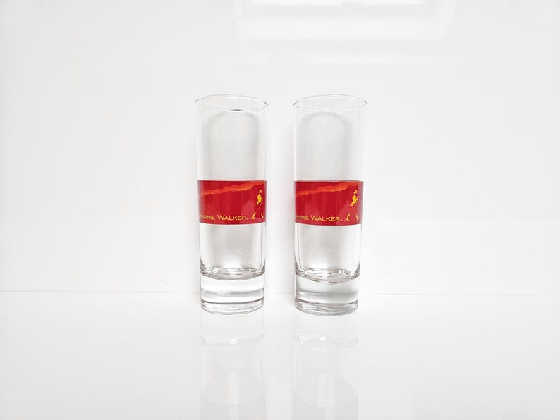 May include: Two clear, tall shot glasses with a red band featuring the text "Johnnie Walker". The glasses are filled with a clear liquid and sit on a white surface.