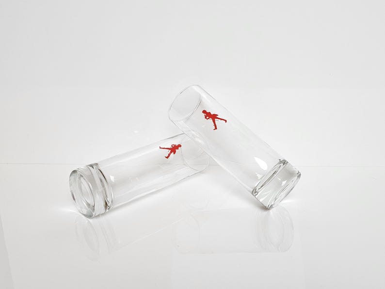 Peut inclure: Deux verres &agrave; boire cylindriques transparents, chacun orn&eacute; d'une silhouette rouge d'une personne. Les verres sont l&eacute;g&egrave;rement inclin&eacute;s, mettant en valeur leur forme et leur design minimaliste. Parfaits pour les boissons.
