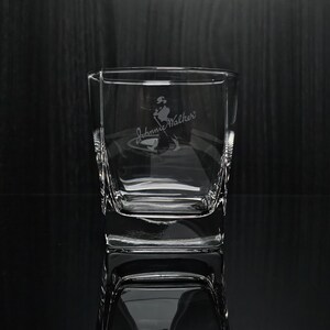 May include: A clear, square-shaped glass with etched "Johnnie Walker" branding and a logo of a man walking. The glass has a thick base and textured sides. The glass is displayed on a reflective surface.