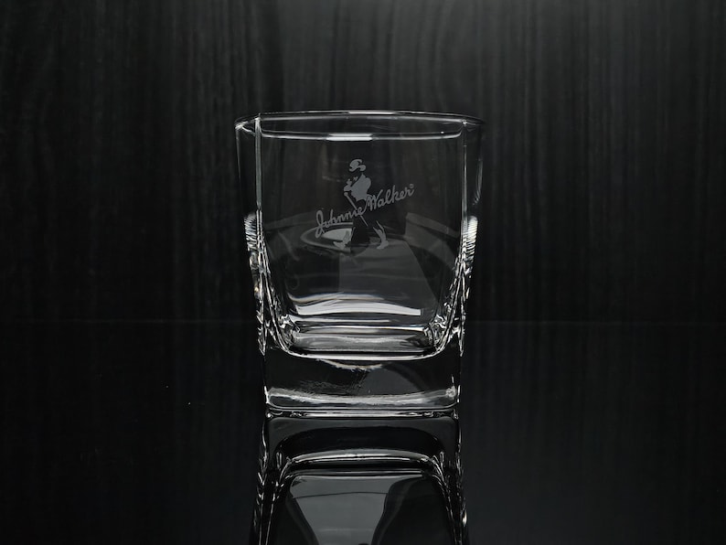 May include: A clear, square-shaped glass with etched "Johnnie Walker" branding and a logo of a man walking. The glass has a thick base and textured sides. The glass is displayed on a reflective surface.