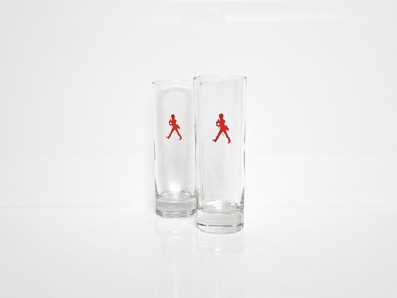 Peut inclure: Deux verres &agrave; boire cylindriques transparents avec un logo rouge repr&eacute;sentant une silhouette d'homme en marche. Les verres sont en verre transparent et sont pos&eacute;s sur un fond blanc.