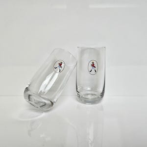 May include: Two clear, cylindrical drinking glasses. Each glass features a circular emblem with a figure in a red coat and black pants. The glasses are positioned on a white surface, one leaning against the other.