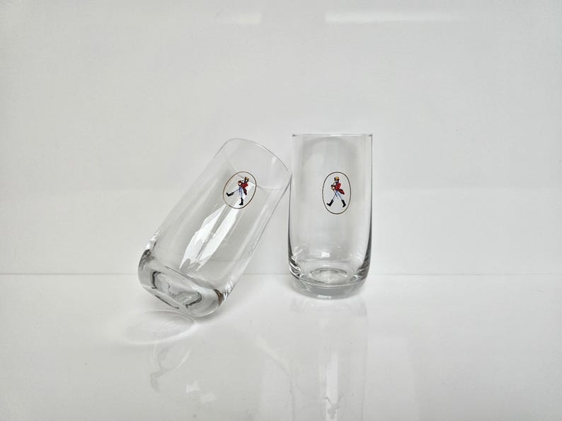 May include: Two clear, cylindrical drinking glasses. Each glass features a circular emblem with a figure in a red coat and black pants. The glasses are positioned on a white surface, one leaning against the other.