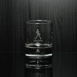 May include: A clear, cylindrical rocks glass with the etched silhouette of a man walking and the words "Johnnie Walker" below. The glass has a thick base and is partially filled with a clear liquid, reflecting the image.