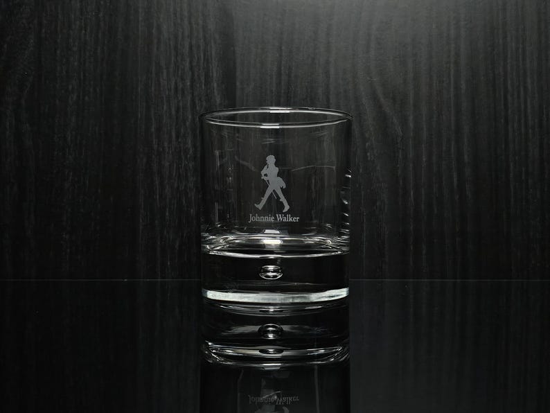May include: A clear, cylindrical rocks glass with the etched silhouette of a man walking and the words "Johnnie Walker" below. The glass has a thick base and is partially filled with a clear liquid, reflecting the image.