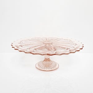 Op de afbeelding: Een roze glazen taartstandaard met een geschulpte rand en een gedetailleerd bloemmotief. De standaard heeft een ronde basis en een voetstuk. De taartstandaard is gemaakt van glas en is lichtroze.