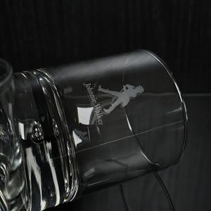 May include: A clear, cylindrical glass with the "Johnnie Walker" logo etched on the side. The logo features a silhouette of a man walking. The glass is partially filled and angled against a dark background.
